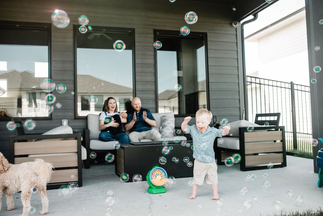 Family watching toddler play with bubble machine on outdoor patio with dog.