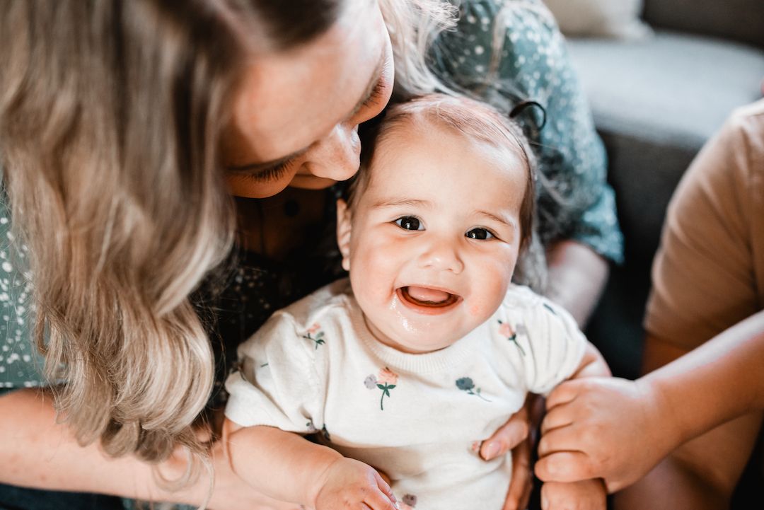 Adorable smiling baby in white onesie being held by parents in a warm home setting.