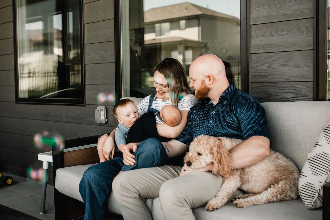 Young family with newborn baby, toddler, and golden dog relaxing on an outdoor porch couch.
