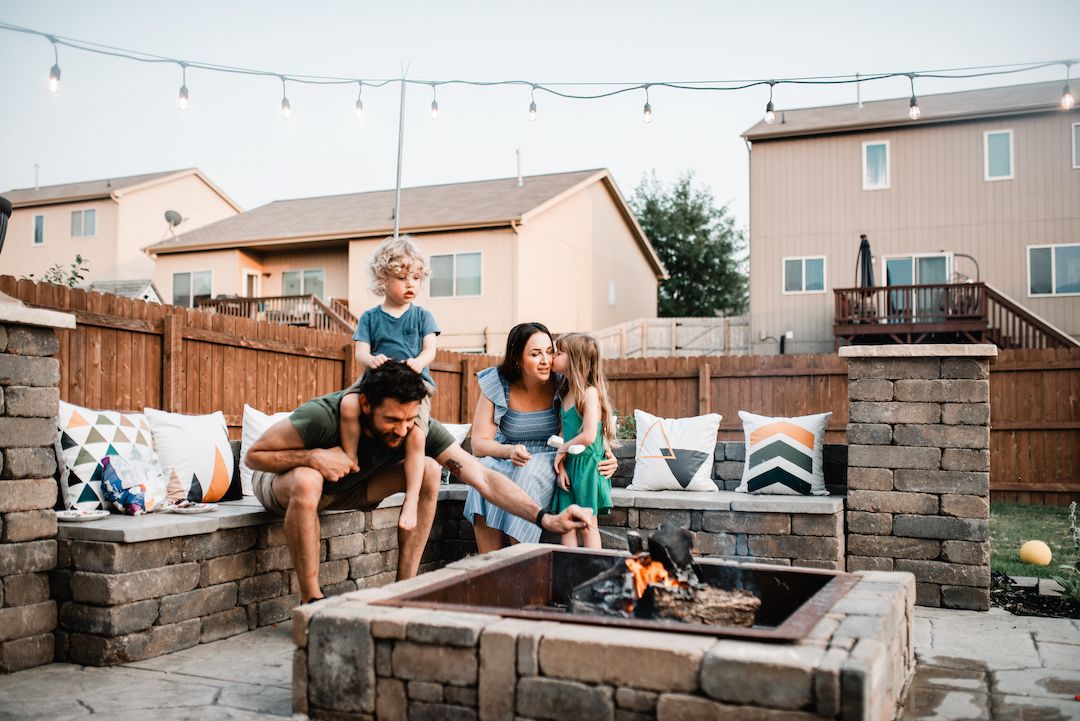 Family of four enjoying a backyard fire pit together at dusk with string lights overhead.
