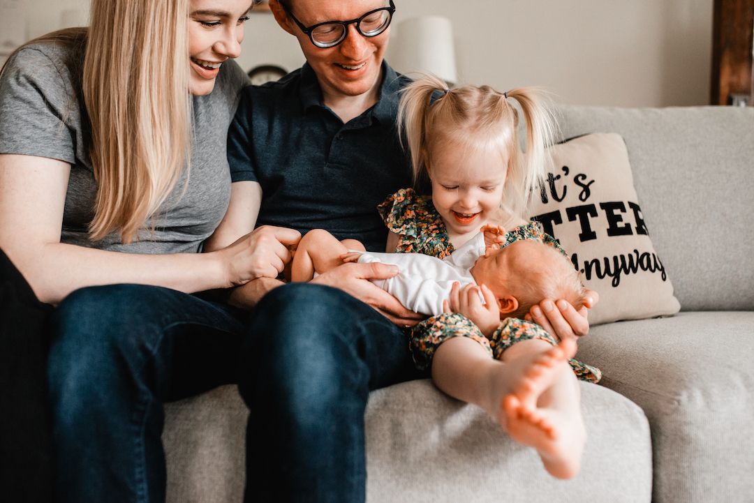 Smiling parents with toddler daughter holding newborn baby on gray sofa.