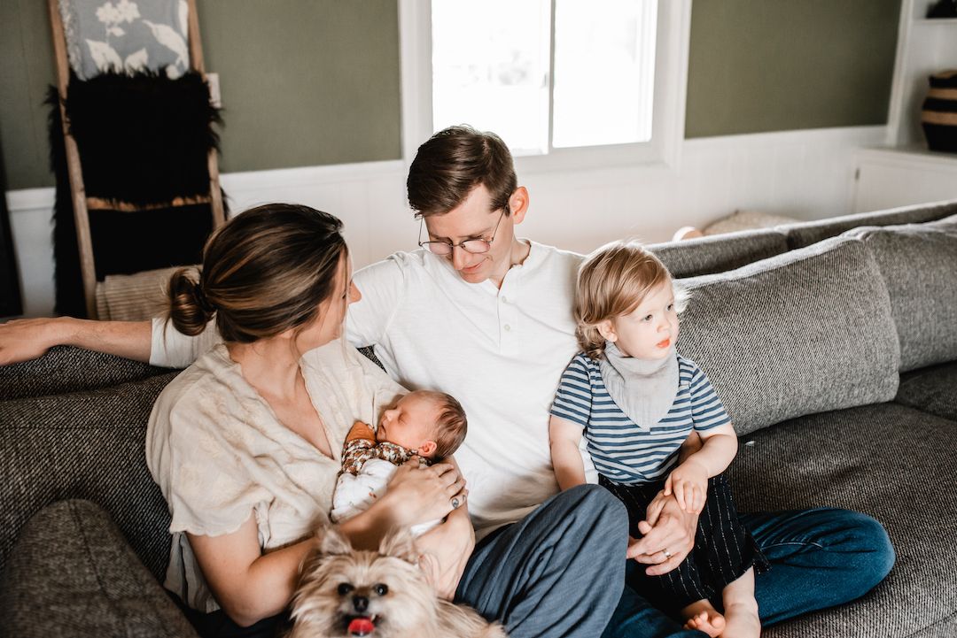 A young family with a newborn baby and toddler sits together on a gray couch with their small dog.