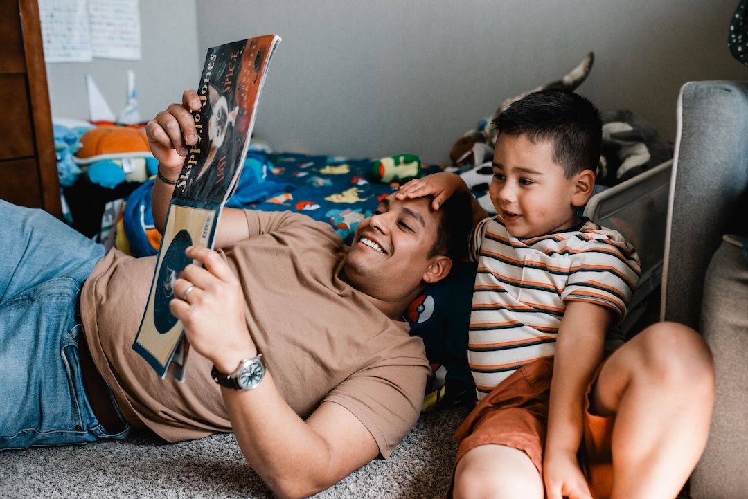Father and young son laughing together while reading a colorful picture book on the floor.