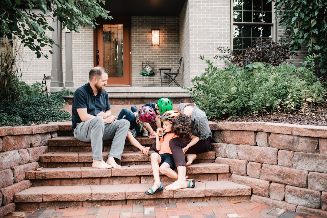 Family of four sitting together on brick steps outside their home in a lush garden setting.