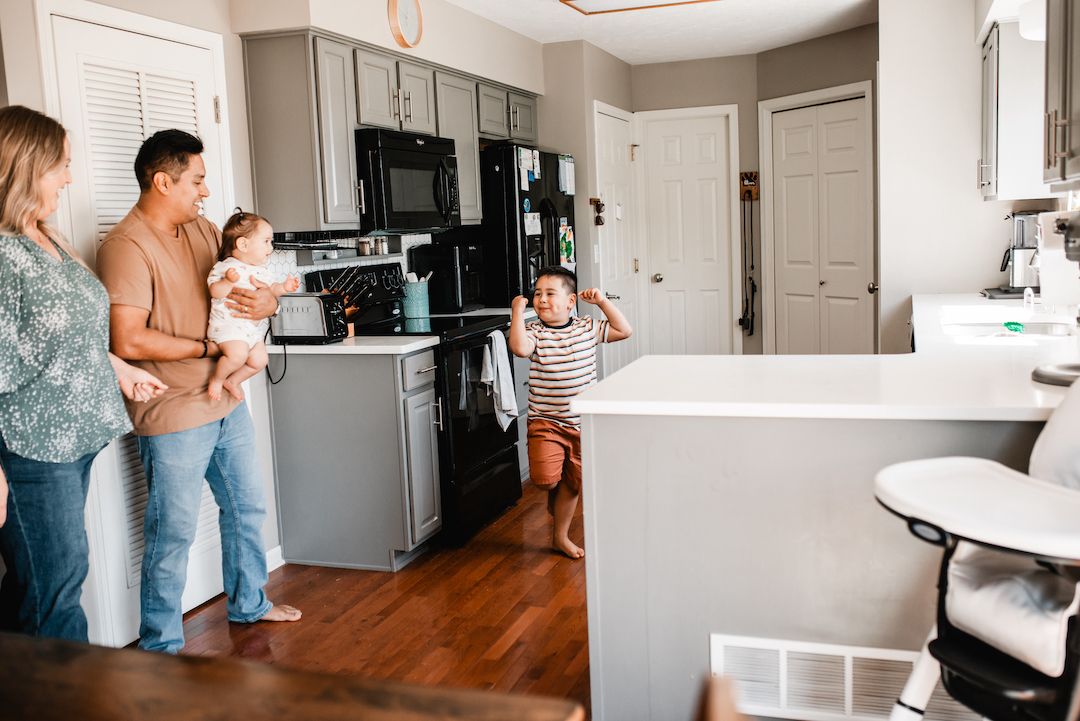 Happy family with baby and young child gathered together in a modern kitchen with gray cabinets.