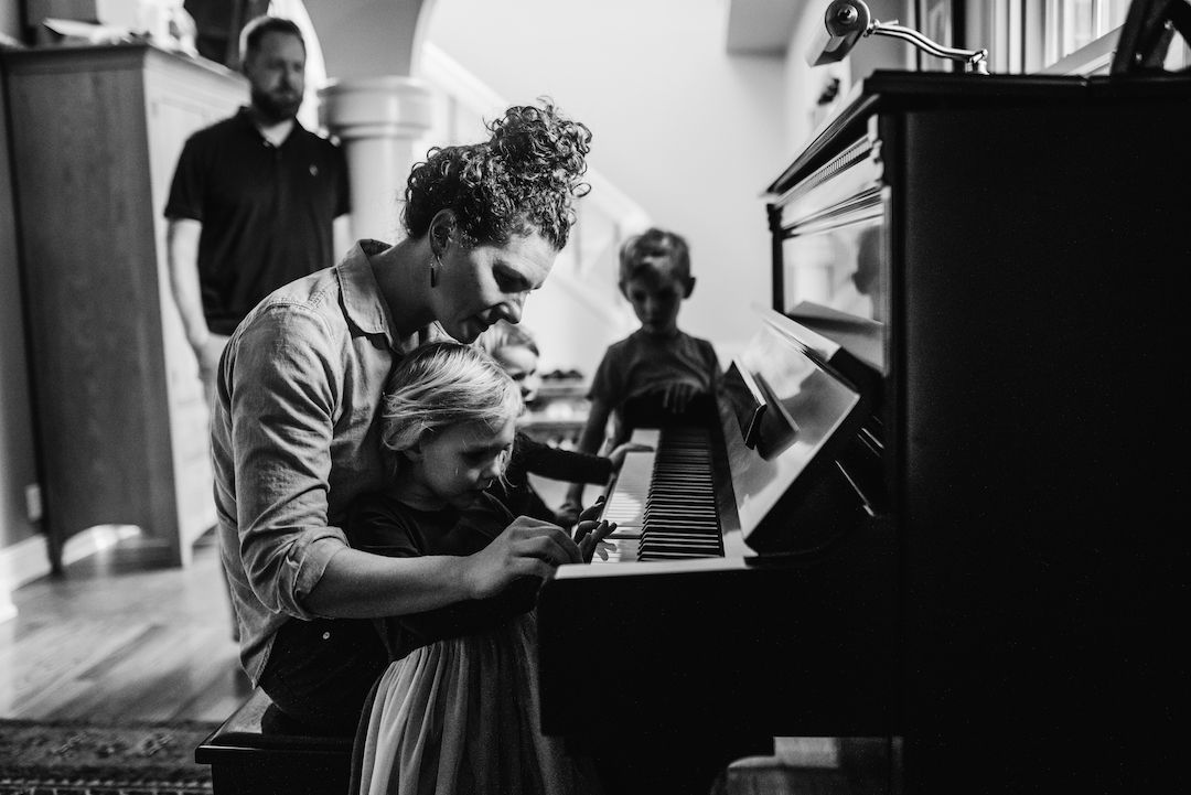 Black and white photo of parent teaching child to play an upright piano in a home setting.