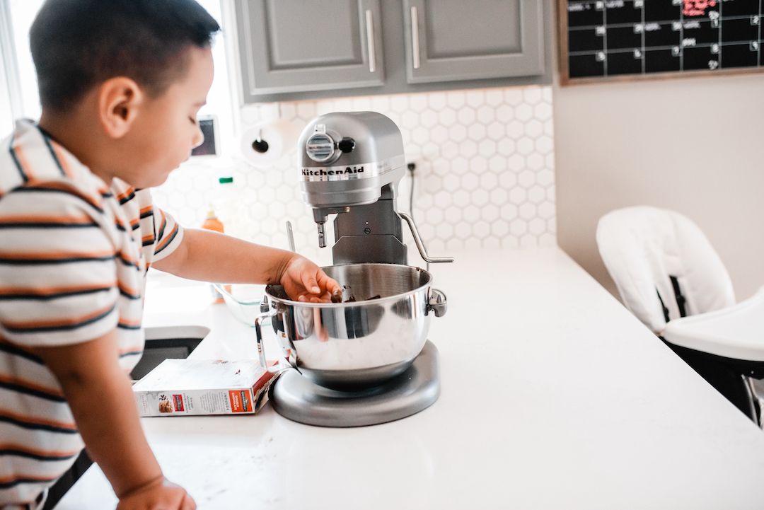Young boy reaching into a KitchenAid stand mixer bowl on a white kitchen counter.