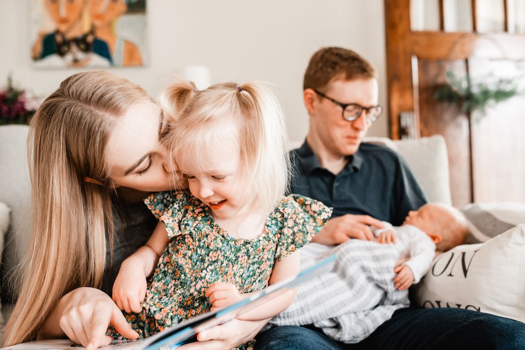 Mother kissing toddler daughter while reading a book as father holds newborn.