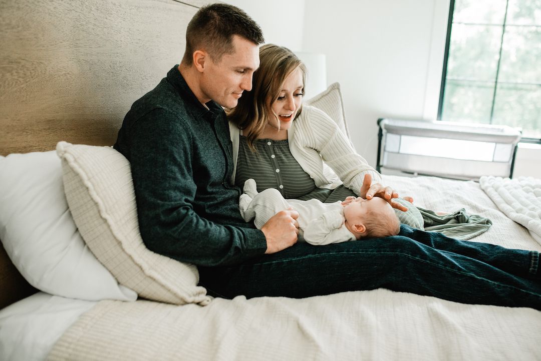 A couple sits on a bed smiling and playing with their newborn baby in a cozy bedroom setting.