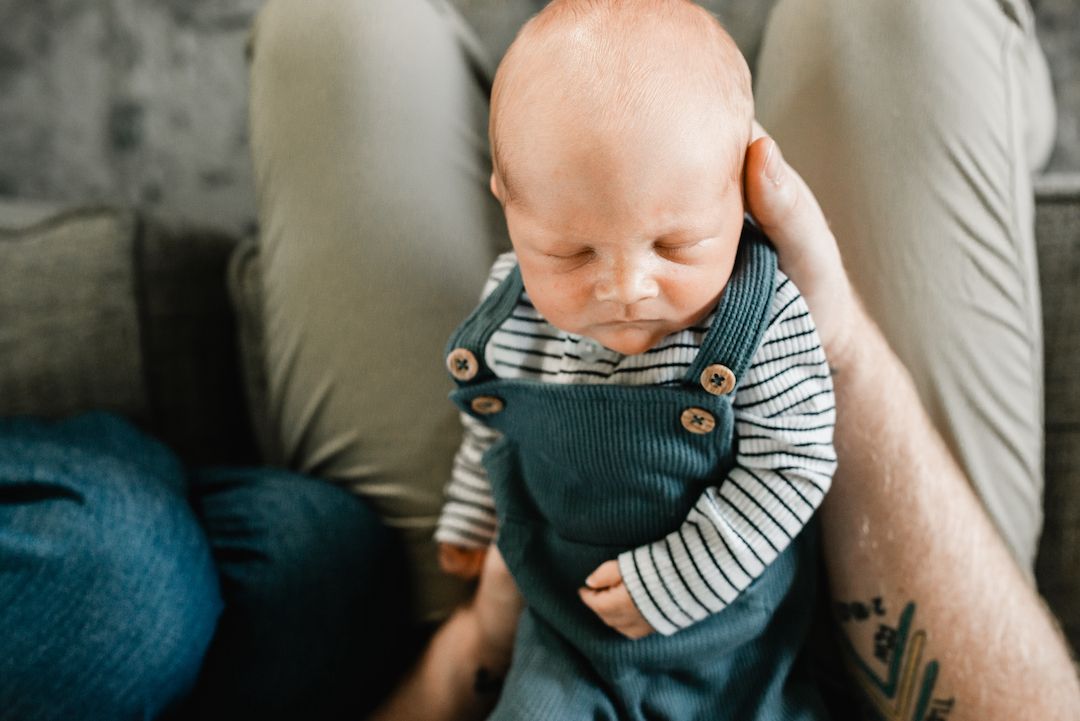 Close-up of a newborn baby in teal overalls and striped shirt cradled in parent's arms.