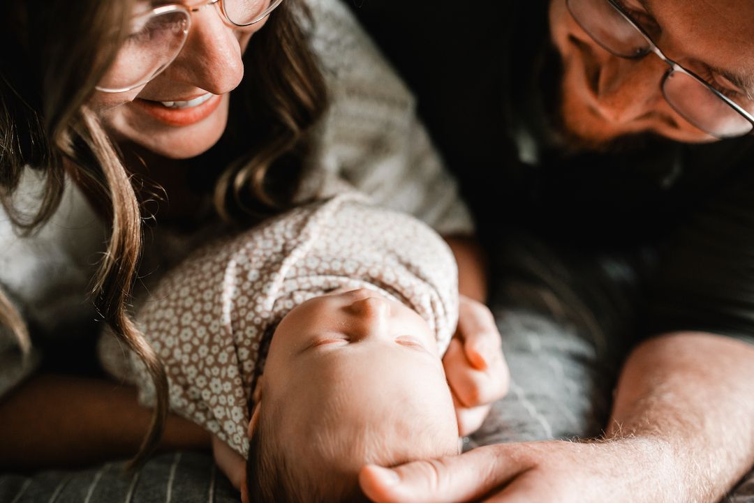 Two parents wearing glasses lovingly gazing at their sleeping newborn baby wrapped in a white floral swaddle.