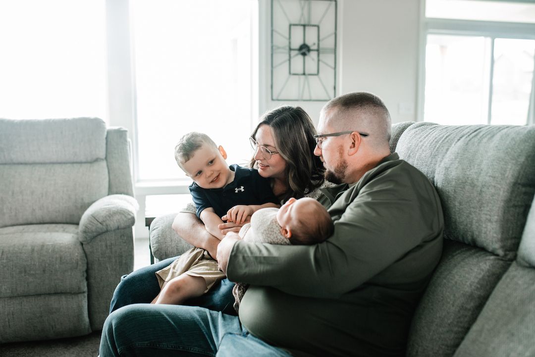 Family of four sitting on a gray couch, parents smiling at toddler and newborn baby in a cozy living room.