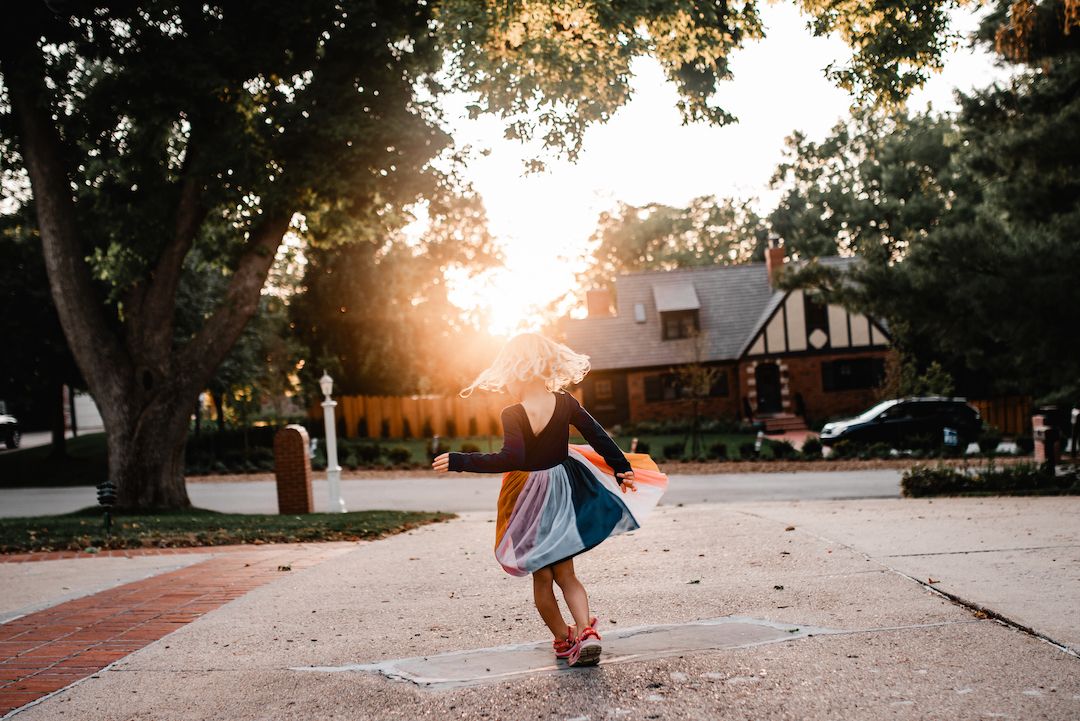 A young girl twirls in a colorful skirt on a sunlit path with golden hour light glowing behind her.