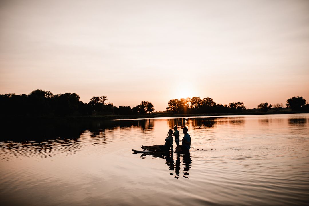 Silhouette of family standing in a lake during a golden sunset with trees reflecting on calm water.