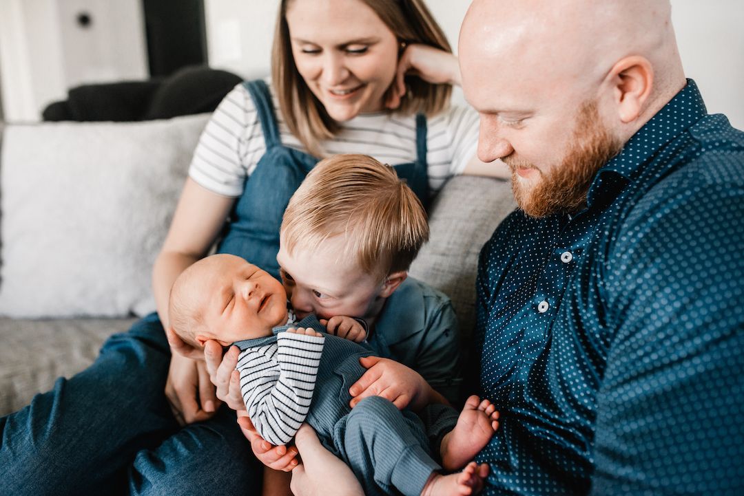 Happy family with red-haired dad, mom, toddler, and newborn baby sitting together on a couch.