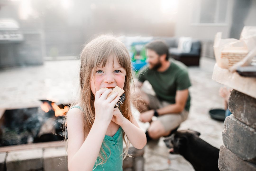 A young girl enjoys a s'more by a backyard fire pit while her father relaxes in the background.