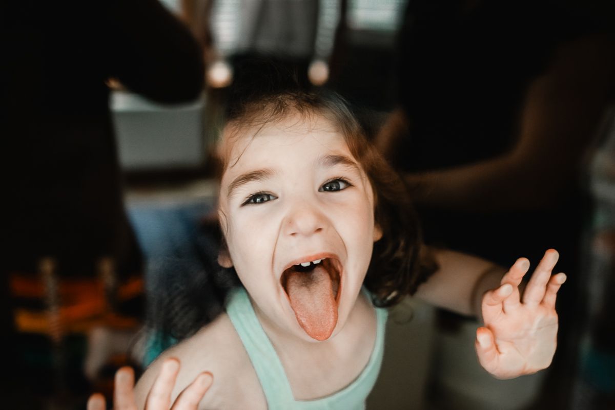 A young girl sticking out her tongue and making a silly face with hands raised, wearing a light blue tank top.