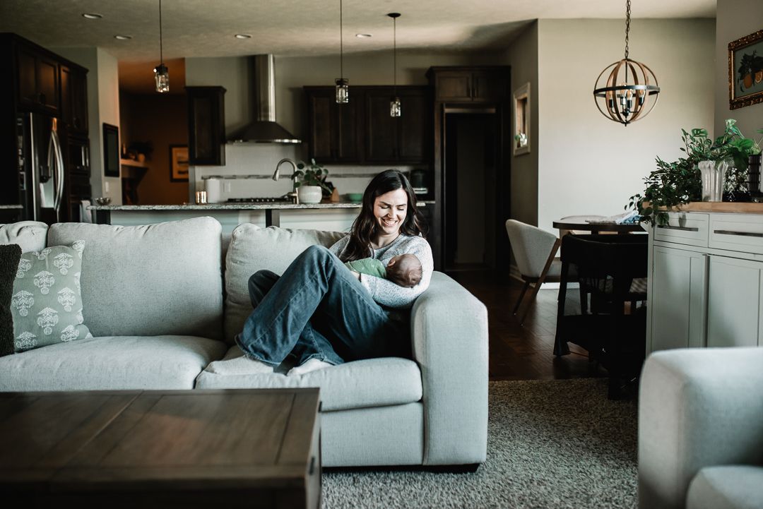 Mother sitting on a gray couch cradling her newborn baby in a modern open-concept living room.