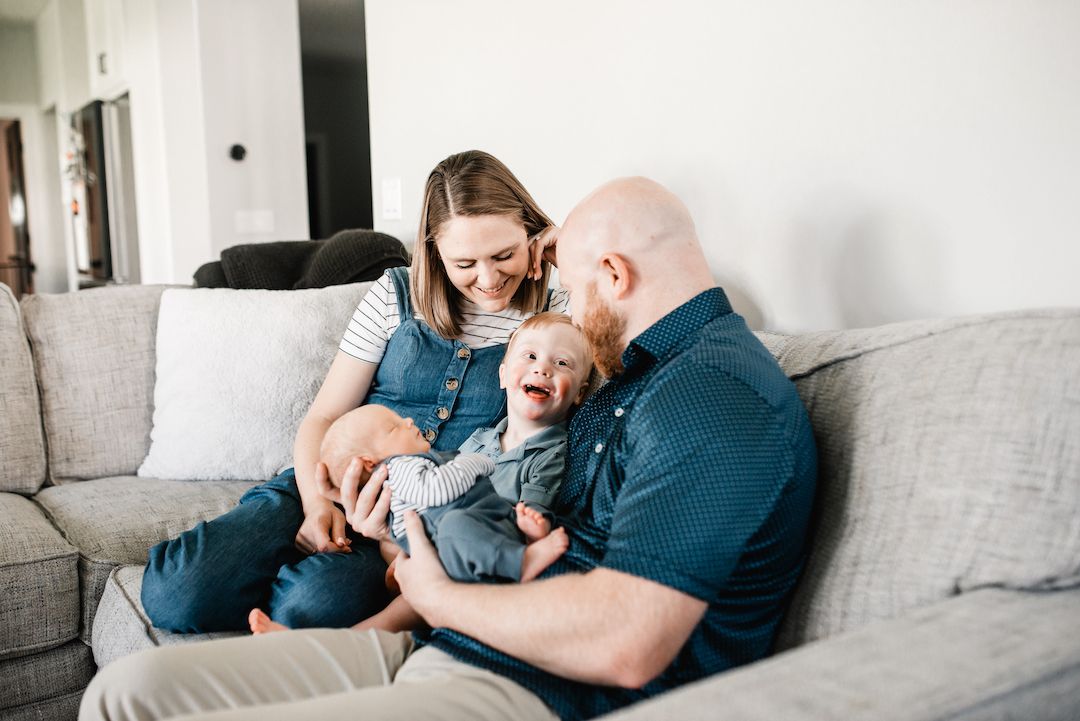 A happy family with a bald father, smiling mother, and laughing baby sitting together on a gray couch at home.
