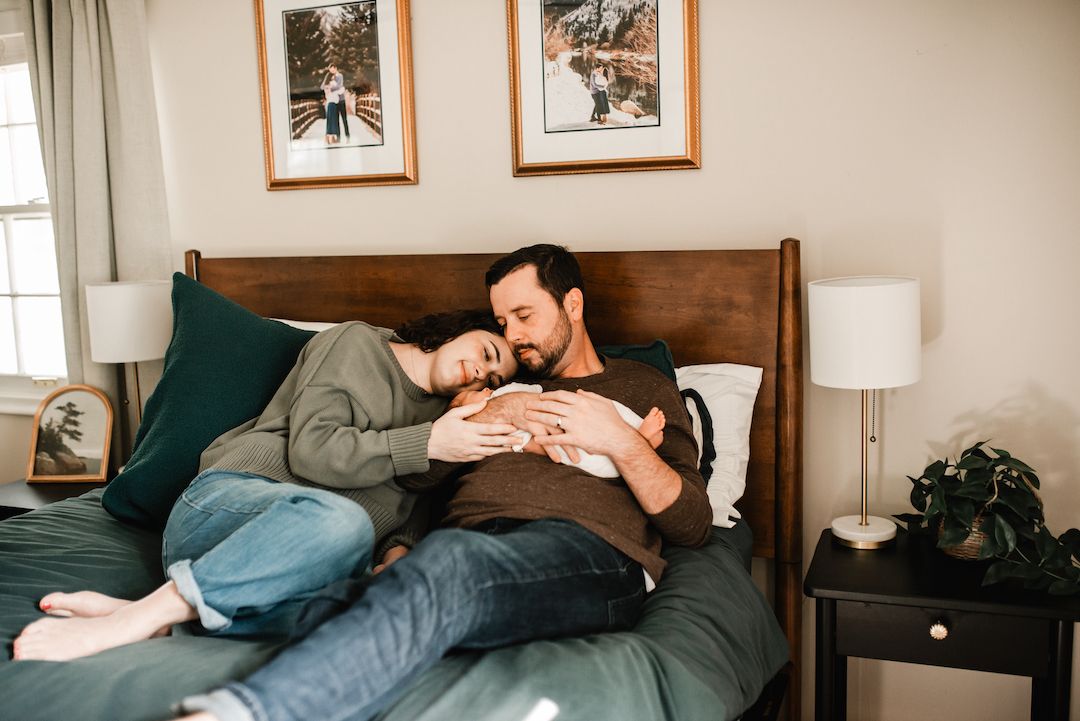 Couple resting on a bed with a newborn baby, sharing a tender family moment in a cozy bedroom.