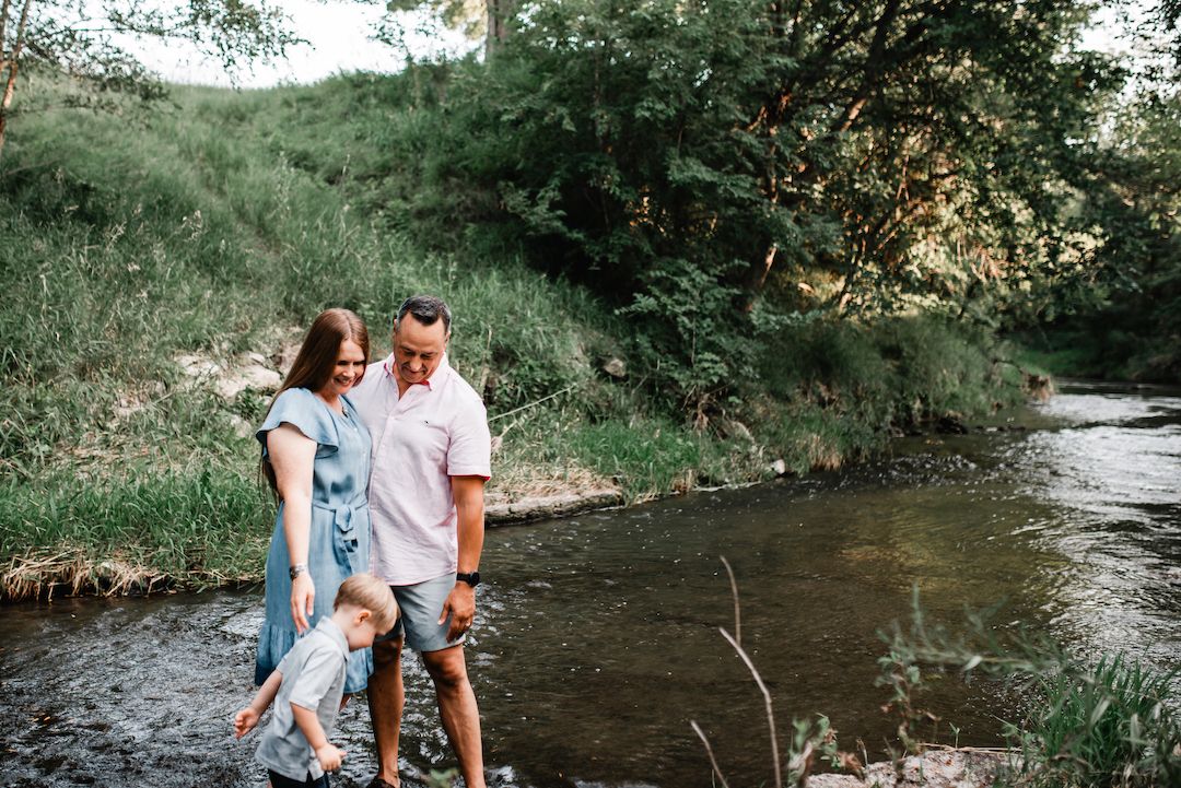 Young family of three wading in a shallow creek surrounded by green summer foliage.