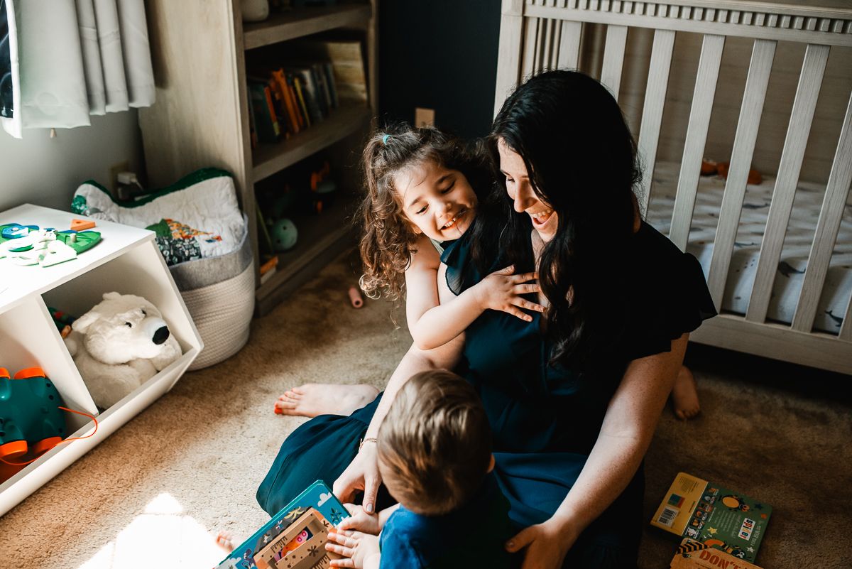 A mother sits on the floor smiling with her two young children in a cozy nursery room filled with books and toys.