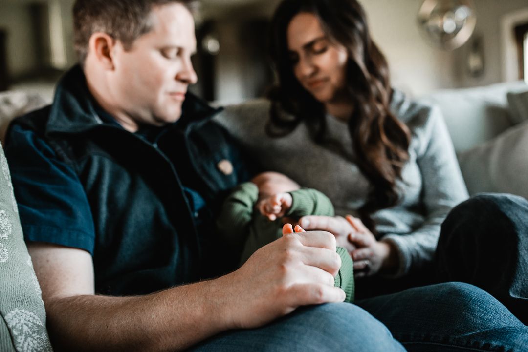 A couple sits on a couch together, lovingly looking at their newborn baby wrapped in green.