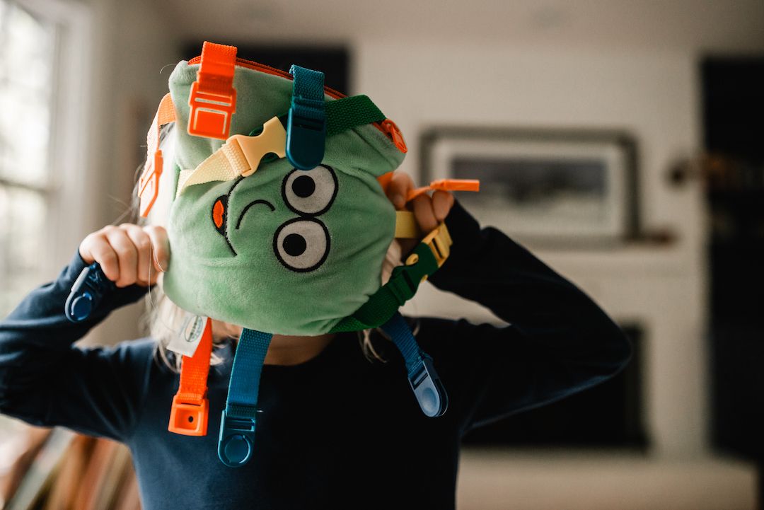 Child wearing a fun green monster backpack covering their face indoors.