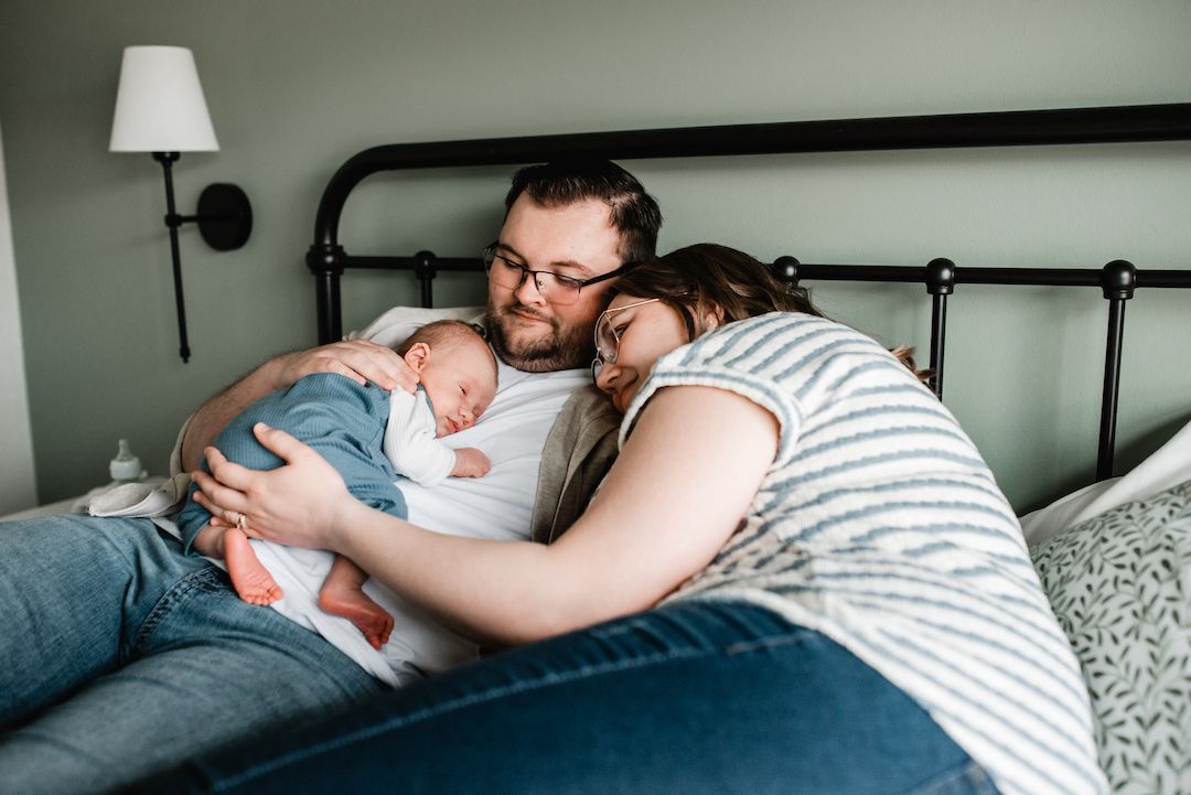 New parents cuddle their sleeping newborn baby together on a bed in a cozy bedroom setting.