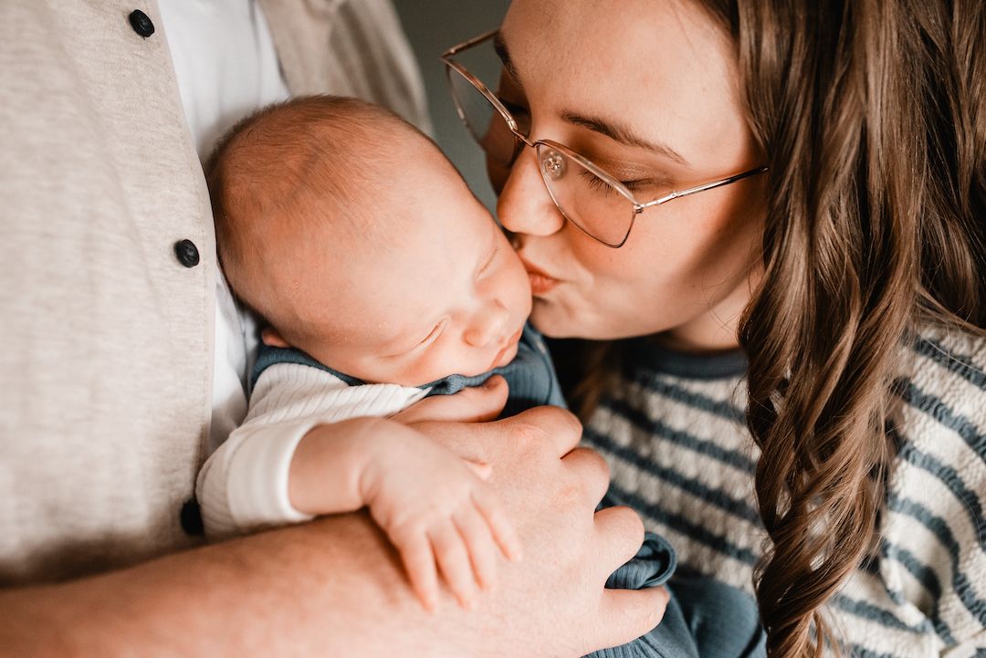 A mother with glasses kisses her newborn baby held in her arms, with a man in a gray shirt beside her.