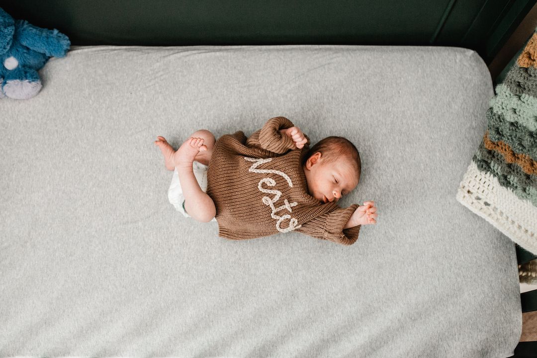 Newborn baby sleeping peacefully in a crib wearing a brown knit sweater with name embroidered on it.