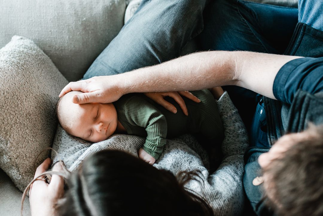 Parents cradling a sleeping newborn baby in a green onesie on a cozy gray couch.