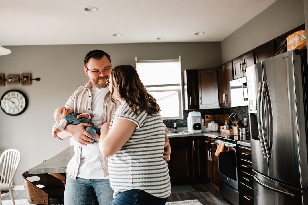 A happy couple dances in their modern kitchen while holding their newborn baby at home.