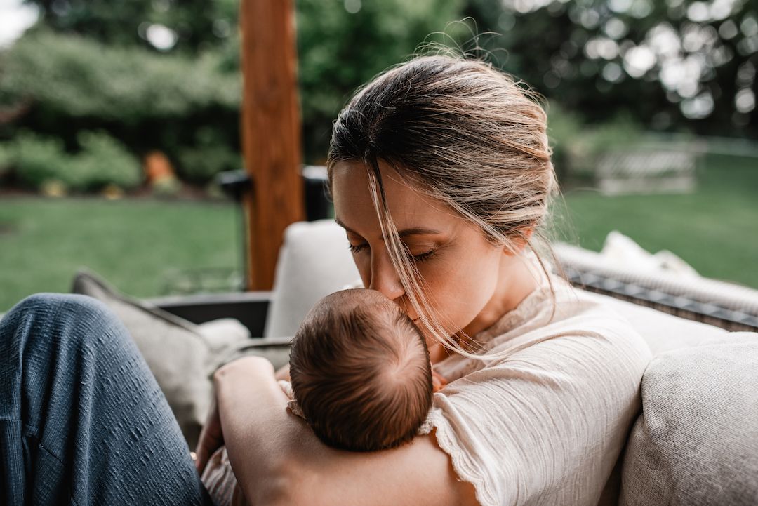 Young mother lovingly kissing her newborn baby while sitting on outdoor patio sofa.