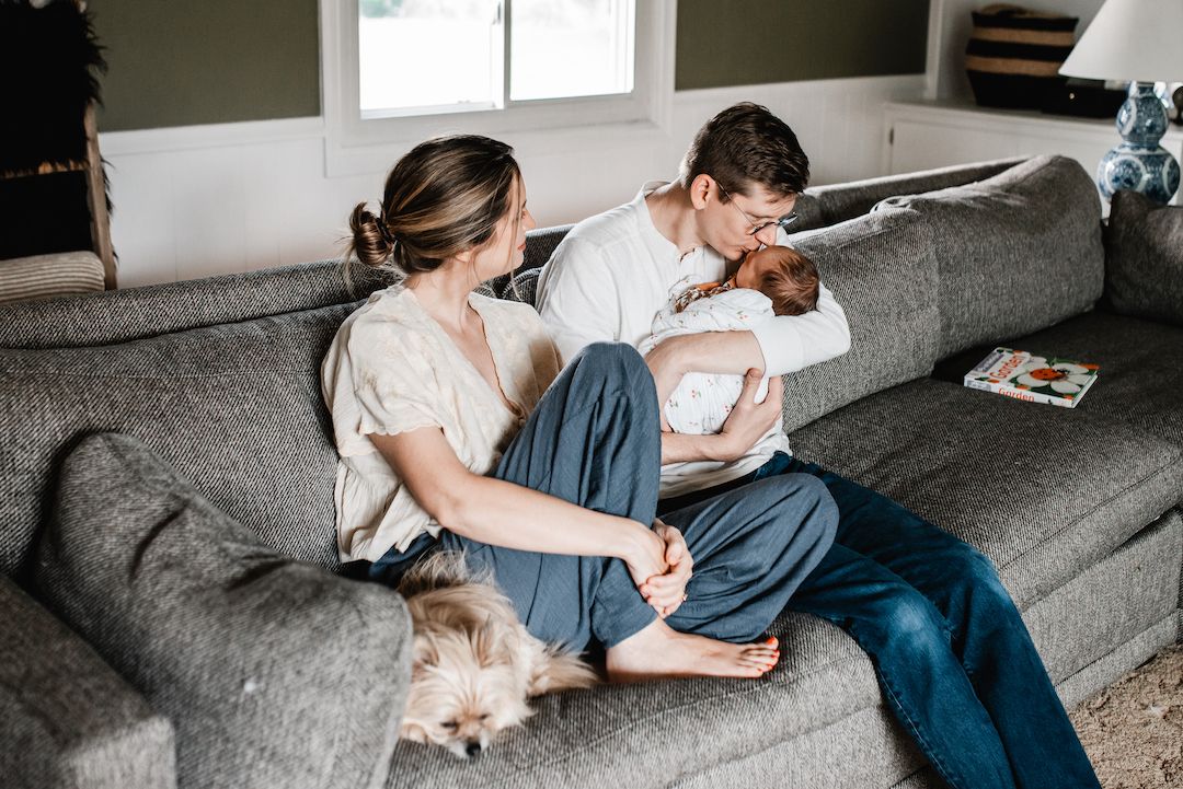Young couple with newborn baby and small dog relaxing together on gray couch in cozy living room.