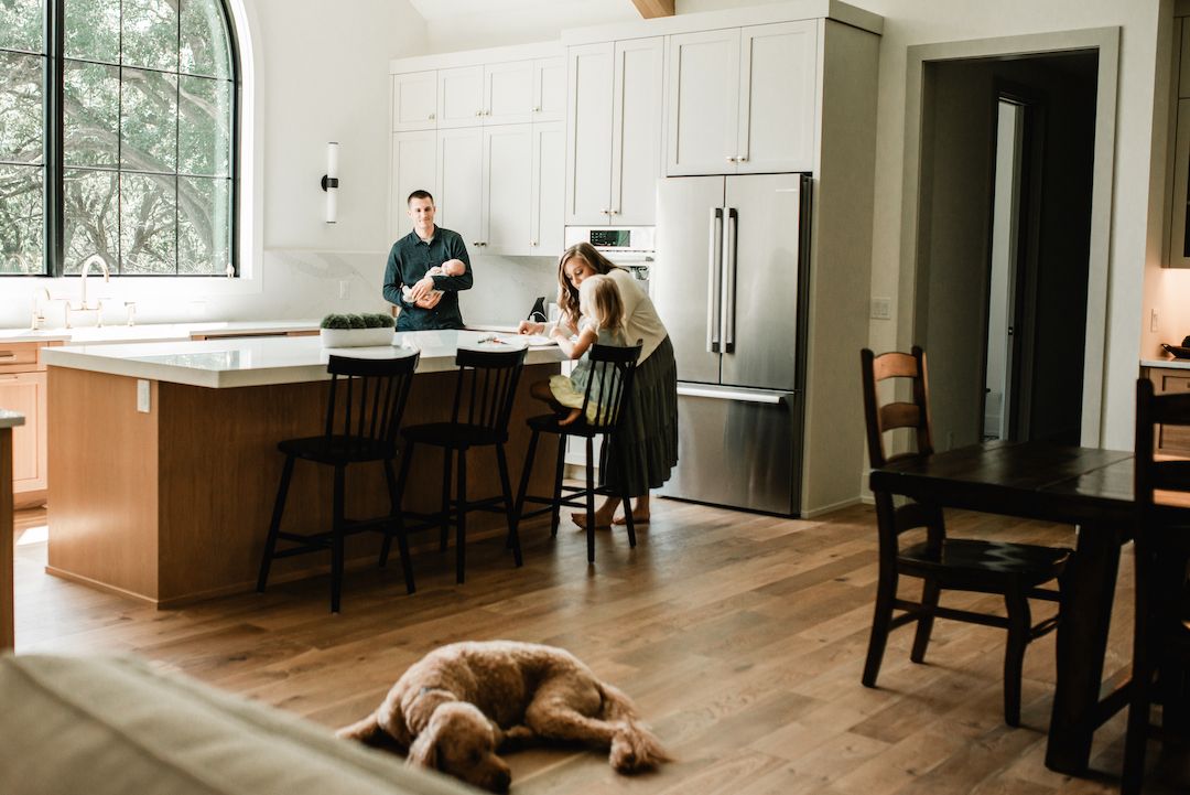 Family in modern kitchen with dog resting on hardwood floor, man and woman at island with bar stools.