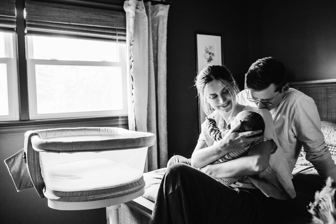 Black and white photo of new parents smiling at their newborn baby beside a bassinet in a cozy bedroom.