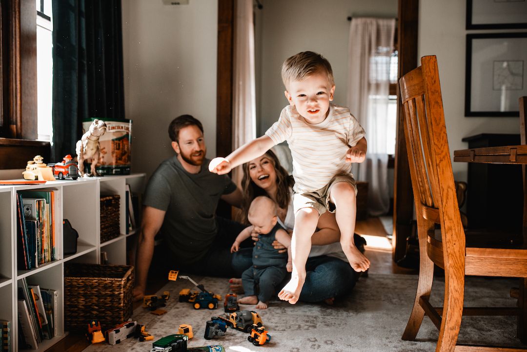 Joyful family playing with toys on floor while toddler leaps up.