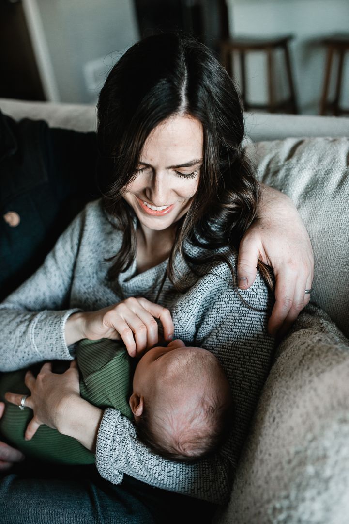 Smiling dark-haired woman in gray sweater cradling a newborn baby on a couch indoors.