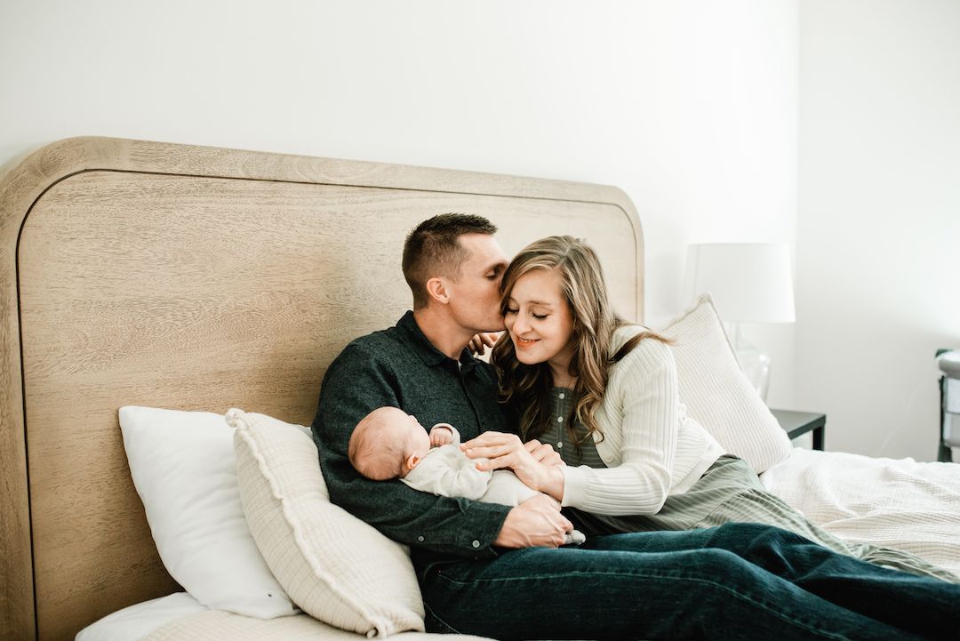 Happy couple sitting on bed admiring their newborn baby in a bright cozy bedroom.