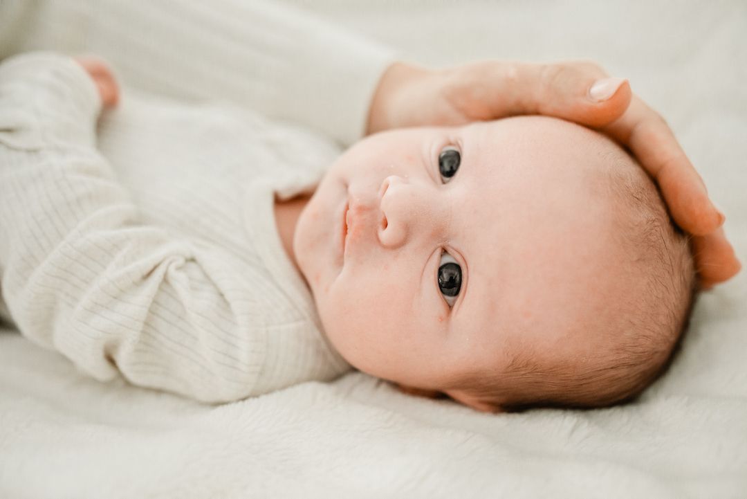 Close-up of a newborn baby in a white onesie lying on a soft blanket with an adult hand on head.