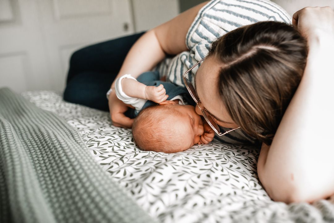 A mother tenderly gazes at her sleeping newborn baby while lying on a bed with a patterned blanket.