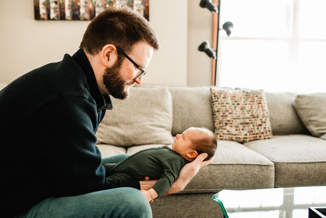 Bearded father with glasses lovingly gazing at his newborn baby on a couch at home.