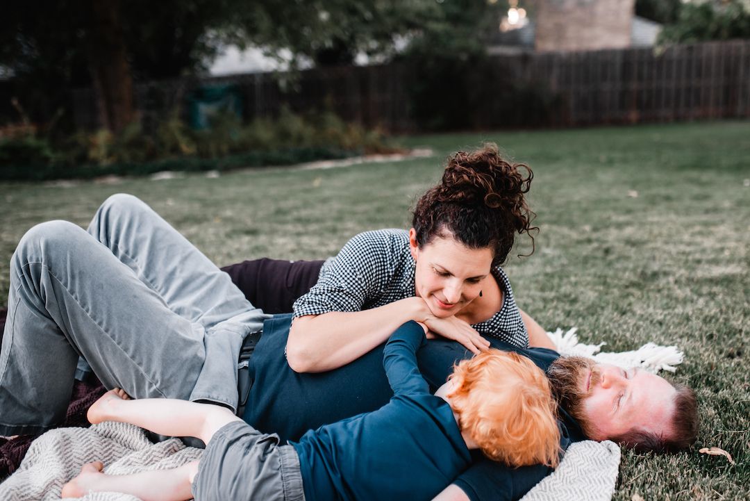 A joyful family of three laughs and plays together on a blanket in the backyard.