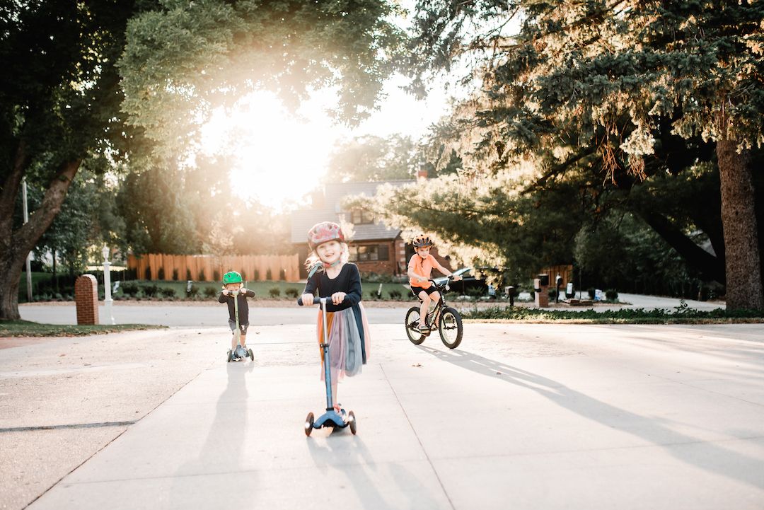 Two children riding a scooter and bike on a sunny tree-lined suburban street.