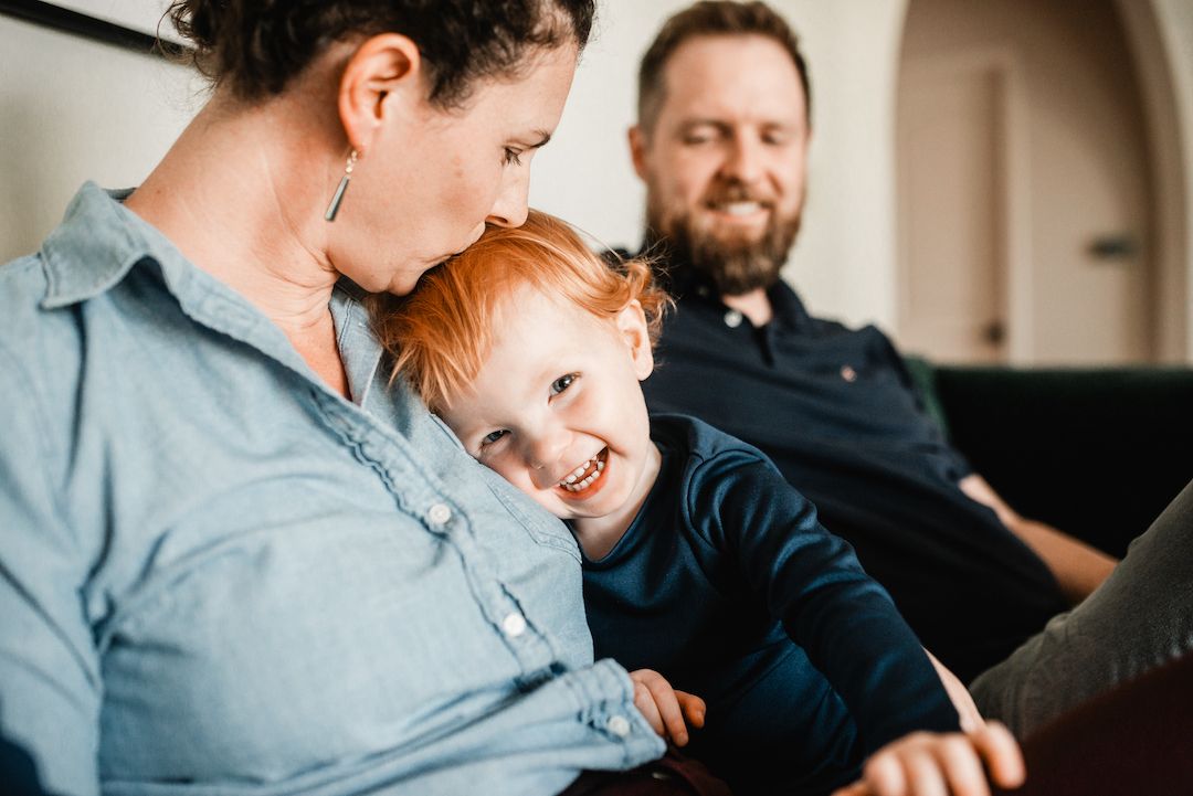 Happy redheaded toddler laughing while snuggling with parents on a comfortable sofa.