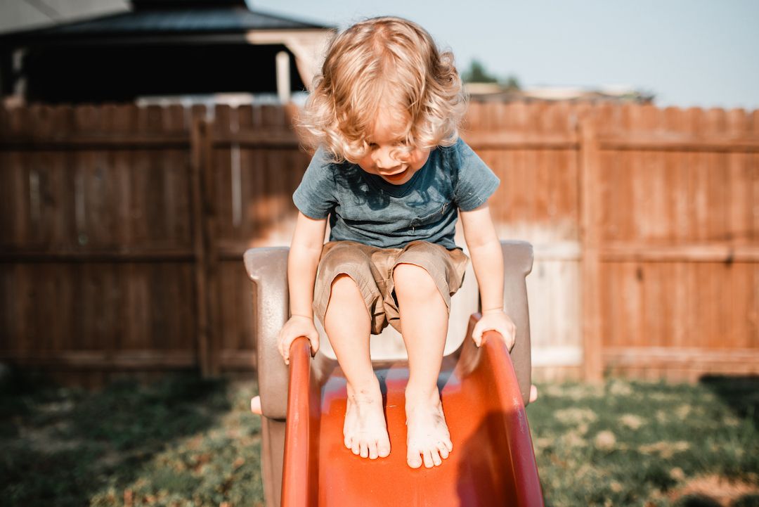 Blonde toddler sitting at the top of a red backyard slide on a sunny day.