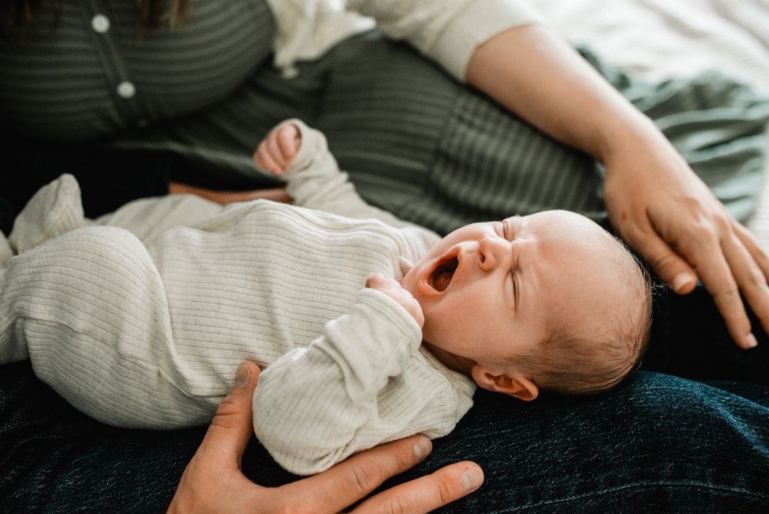 Newborn baby yawning while lying in parents arms on bed.