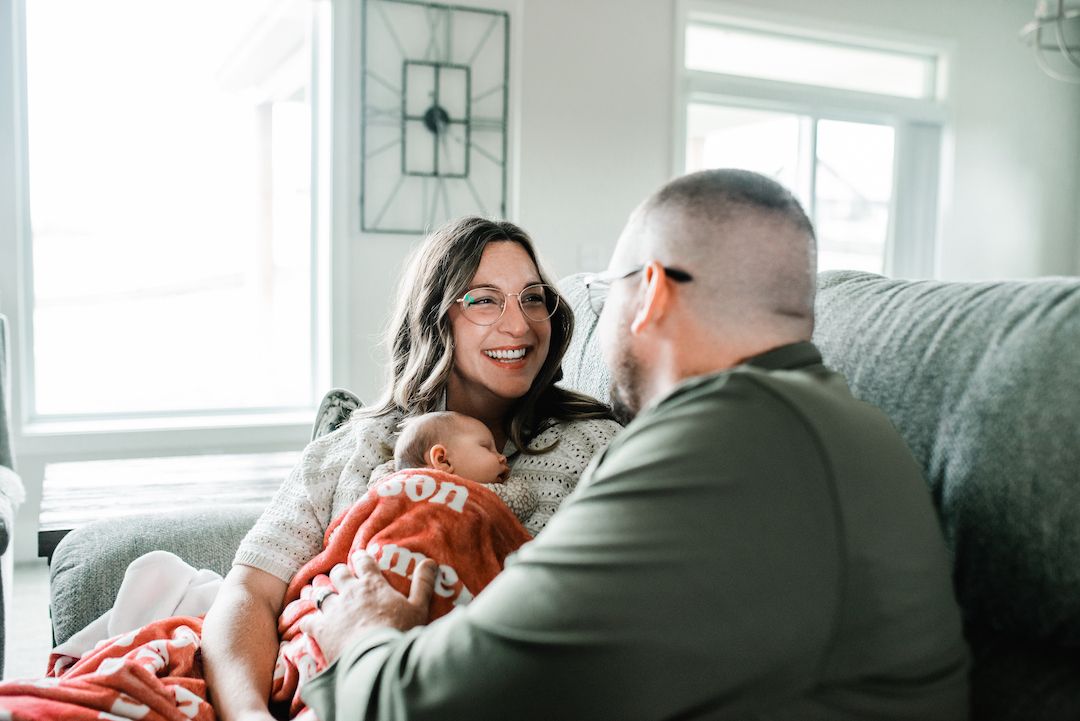 A smiling woman holds a newborn baby on a couch while a man looks on in a bright living room.