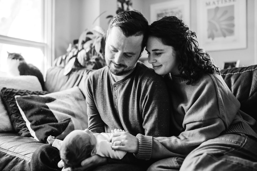Black and white photo of couple tenderly holding newborn baby together on couch at home.